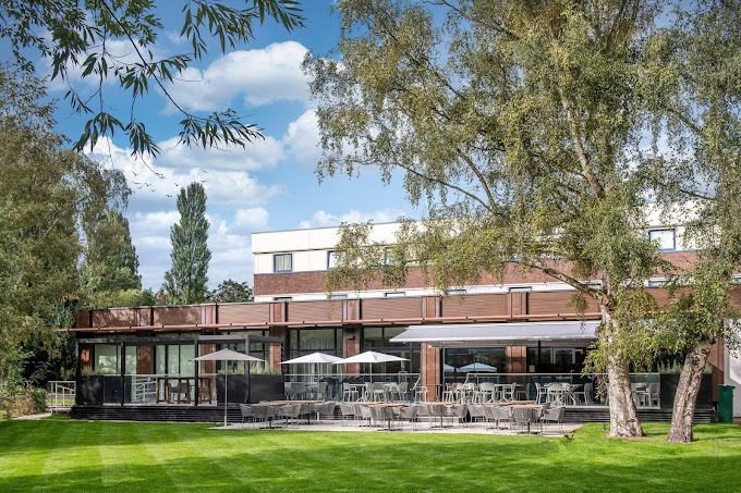 Holiday Inn Shepperton exterior with an outdoor patio, umbrellas, and lawn surrounded by trees.